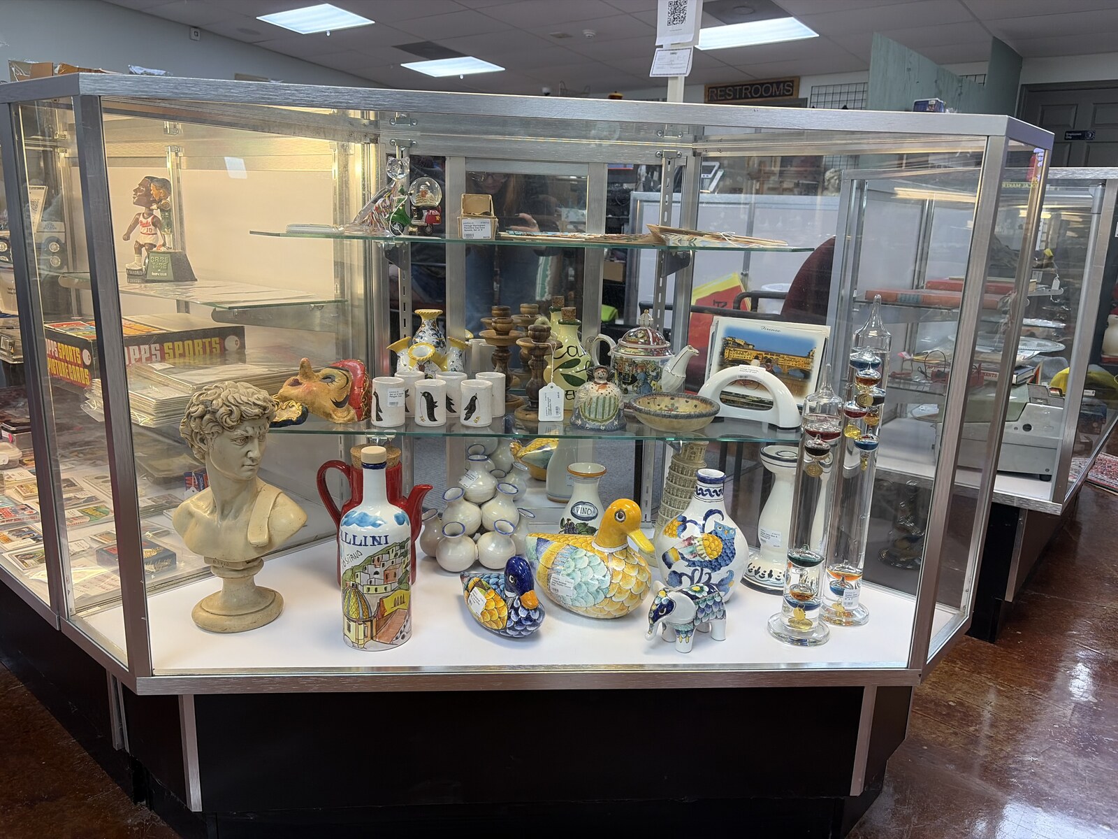 Treasures from Tuscany display case inside Triangle Trading Post with hand-painted ceramic ducks, an owl vase, a classical bust, a Leaning Tower of Pisa figure, and glass Galileo thermometers