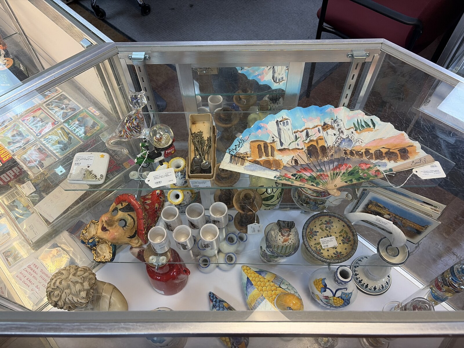 Top-down view into the Treasures from Tuscany case showing a hand-painted Assisi fan, a glass cat figurine, a Vespa snow globe, souvenir spoons, a painted mask, and hand-painted Italian pottery